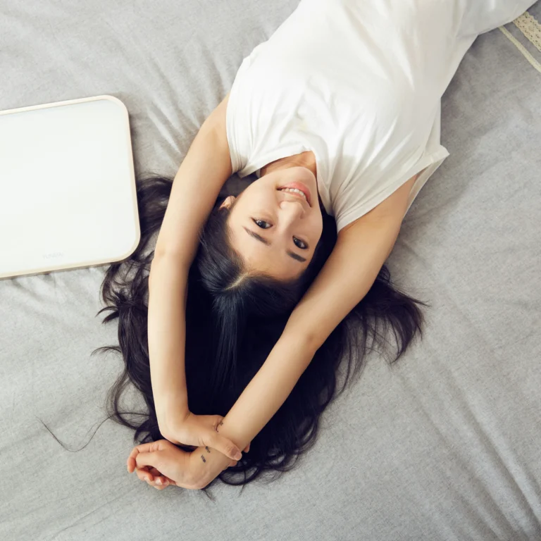 Smiling woman relaxing on a soft gray bed sheet, arms stretched overhead, enjoying comfort and relaxation.
